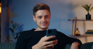 Evdeki kanepede otururken akıllı telefonunu kullanan heyecanlı bir esmer adam. Yakışıklı milenyum erkeğinin görünüş ve kaydırmalı telefon ekranı. Boş zaman kavramı..