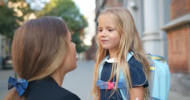 Zblízka pohled na malé roztomilé dítě objímající svou matku před odchodem ze školy na ulici. Mladá atraktivní matka mluví a mává své dceři, že běží do třídy v dopoledních hodinách. — Stock video