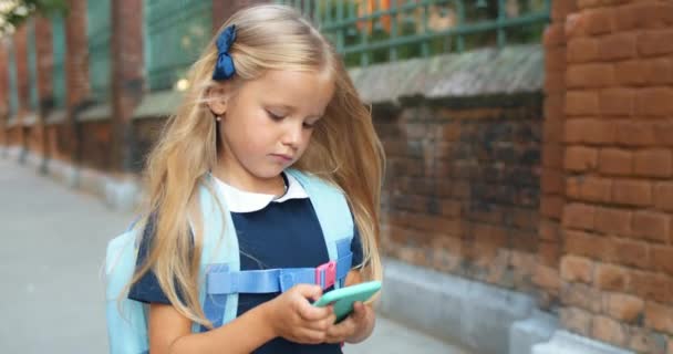 Entzückende Schülerin mit modernem Smartphone beim Gehen. Hübsches Kind mit langen blonden Haaren und Rucksack, das zur Schule geht, während es guckt und den Handybildschirm berührt. Konzept der igeneration. — Stockvideo