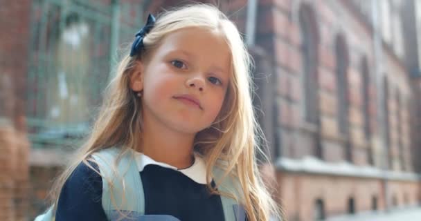 Csinos kislány portréja, ahogy a kamerába néz. Közelkép egy imádnivaló, hosszú, szőke hajú kölyökről, aki pózol, miközben az utcán áll. Fogalom a gyermekek és az oktatás. Régi épület a háttérben. — Stock videók