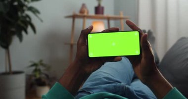 Elinde yeşil ekran akıllı telefonu olan adam kanepede yatay peyzaj modunda yatarken keçeli kalemler takıyor. Afrikalı adam ekrana bakarken video izliyor..