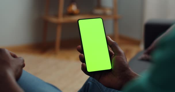 Sopra la spalla vista di uomo afro americano seduto sul divano e tenendo smartphone con finto schermo in mano. Concetto di chiave cromatica e schermo verde. Accogliente sfondo casa . — Video Stock