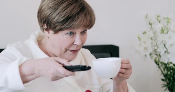 Primer plano de la anciana encendiendo la televisión y sosteniendo la taza mientras está sentada en la cama. Mujer madura sonriente mirando la pantalla de televisión mientras bebe café y disfruta de buenos días. Concepto de relax . — Vídeo de stock