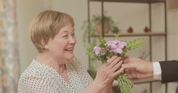 Erkeklerin yaşlı güzel bir kadına el sallamalarının yan görüntüsü. Mutlu emekli bayan çiçek alıyor, kameraya gülümsüyor. İnsanlar ve olumlu duygular kavramı. — Stok video