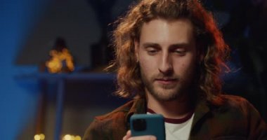 Milenyum sakallı bir adamın akıllı telefon kullanıp yatay moduna geçişini yakından izle. Uzun saçlı ve küpeli hippi evde kanepede otururken daha az video izliyor..