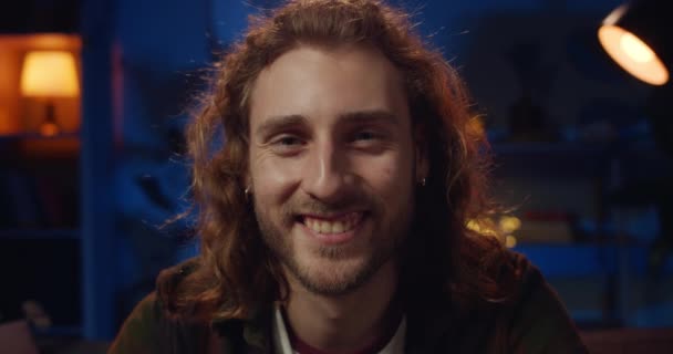 Portret van een jonge hipster met een baard die glimlacht terwijl hij naar de camera kijkt. Close-up zicht van vrolijke langharige duizendjarige man poseren. Concept van emoties.Neon en lamp licht op de achtergrond. — Stockvideo