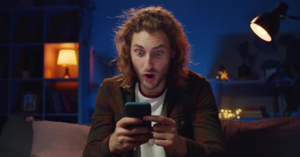 Een jonge hipster die ja zegt en lacht terwijl hij goed nieuws heeft. Knappe langharige man die wint terwijl hij zijn smartphone gebruikt en thuis op de bank zit. Concept van succes. — Stockvideo