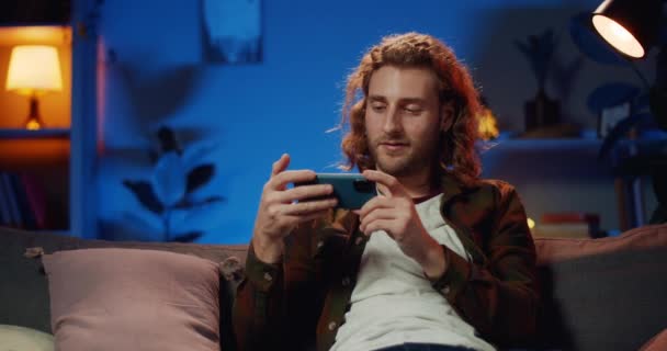 Jonge hipster man roterende smartphone scherm naar landschap horizontale modus en comfortabel zitten op sofa.Bearded man glimlachen tijdens het bekijken van video. Achtergrondinformatie. — Stockvideo