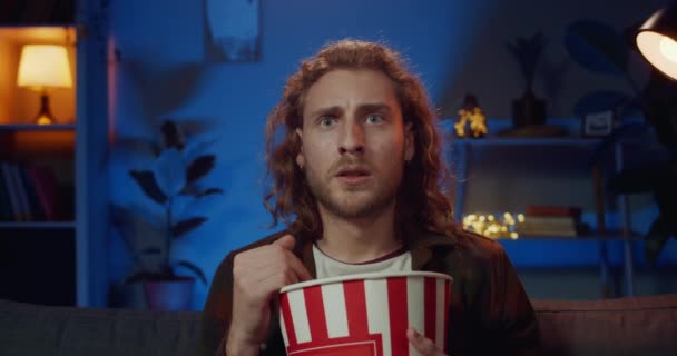 Mladý muž s dlouhými vlasy reagující na děsivou maminku při sledování thrilleru doma. Pohledný muž drží popcorn bowl, zatímco sedí na gauči před televizí. Pojetí emocí. — Stock video