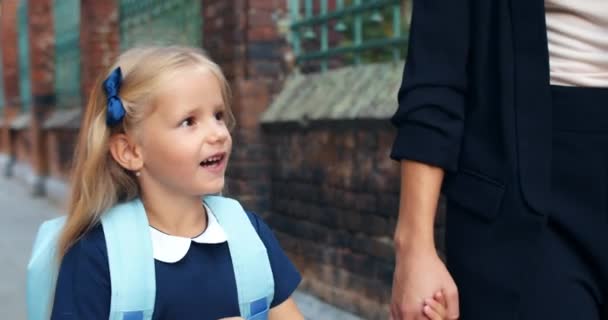 Vue rapprochée d'une jolie écolière aux cheveux blonds marchant et parlant avec sa mère. Doux enfant portant l'uniforme de l'école tenant sa main de mamans sur leur chemin dans la matinée. . — Video