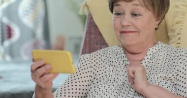 Sandalyede otururken modern cep telefonu kullanan mutlu kadın emekliler. Yaşlı, güzel bir kadın gülümsüyor ve telefon ekranına bakıyor. İnsan ve teknoloji kavramı.