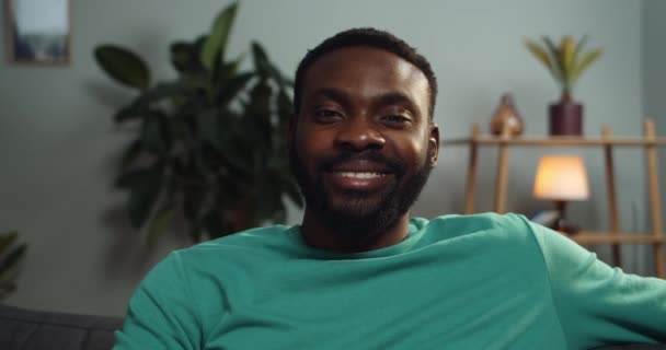 Vista de perto de cara alegre sentado no sofá e sorrindo. Retrato de homem afro-americano olhando e posando para a câmera. Conceito de pessoas e emoções. Fundo de casa . — Vídeo de Stock
