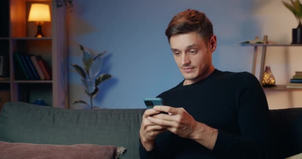 Νεαρός όμορφος άντρας βγάζει σοκαριστικά νέα ενώ χρησιμοποιεί το smartphone του. Χαρούμενος τύπος στα 30 κάνει έκπληξη στο πρόσωπο και τα μεγάλα μάτια ενώ κοιτάζει την οθόνη του τηλεφώνου και κάθεται στον καναπέ στο σπίτι. — Αρχείο Βίντεο