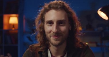 Sakallı, uzun saçlı ve evde kameraya bakan mutlu bir adamın portresi. Küpeli, gülümseyen hippi adamın yakın görüntüsü. Duygu kavramı. Arka planda lamba ışıkları.