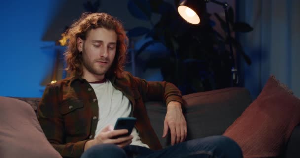 Ung stilig kille som tittar och vidrör skärmen när du använder smartphone. Millennial man med långt hår ler medan du surfar på internet och sitter på soffan hemma. Begreppet fritid. — Stockvideo