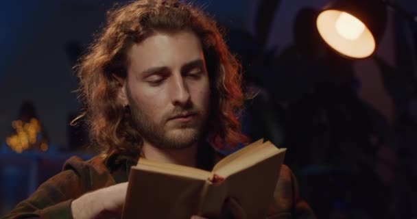 Vista de cerca de inteligente hombre milenario leyendo en la luz de la lámpara mientras se ve somnoliento. Hombre guapo con libro volteando páginas mientras bosteza y se siente cansado. Concepto de ocio . — Vídeo de stock