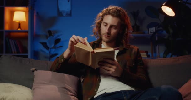 Homem bonito esperto novo que gira páginas do livro ao passar o tempo livre em casa na noite. Millennial cara com cabelo longo leitura literatura interessante enquanto sentado no sofá . — Vídeo de Stock