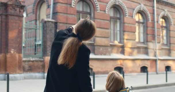 Πλάτη άποψη της μητέρας στέκεται με το μικρό της παιδί χέρι στο χέρι στο δρόμο. Γυναίκα μιλάει με την κόρη της με τσάντα ενώ δείχνει το κτίριο του σχολείου. Έξω. Έννοια της επιστροφής στο σχολείο. — Αρχείο Βίντεο