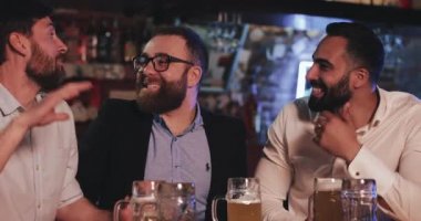 Londra İngiltere - 19 Nisan 2019: Mutlu sakallı adamlar barda birlikte vakit geçirirken keyifleri yerinde. Erkek arkadaşların bira içerken gülüp konuşmalarını ve tezahürat etmelerini yakından izleyin..