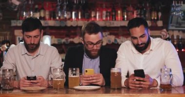 Londra İngiltere - 19 Nisan 2019: İşten sonra bira barında oturan üç adam akıllı telefonlarını kullanıyor. Sakallı adam iş arkadaşlarına cep telefonundan şok edici haberler veriyor ve şaşırmış görünüyorlar..