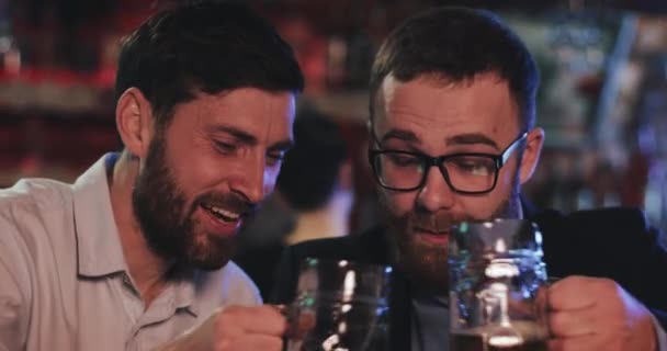 Лондон, Великобритания - 19 апреля 2019 года: закройте глаза на мужчин в хорошем настроении, облизывающих кружки пива во время отдыха после работы. Друзья-мужчины улыбаются и пьют пиво холодного разлива, прекрасно проводя время в пабе . — стоковое видео