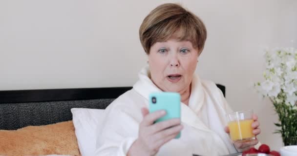 Ältere Hausbesetzerinnen schauen geschockt, während sie die neuesten Nachrichten auf ihrem Smartphone lesen. Reife Frau hält Glas Saft in der Hand und benutzt Handy beim Frühstück im Bett. — Stockvideo