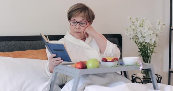 Mujer madura concentrada en gafas leyendo novela durante el desayuno en la cama. anciana vistiendo casa roba pasar tiempo con libro mientras está sentado con bandeja llena de comida en las piernas . — Vídeos de Stock