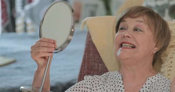 Senior beautiful woman checking her look and smiling in front of mirror. Elderly female retired doing face message and touching face while sitting in chair at home. Concept of beauty. — Stock Video