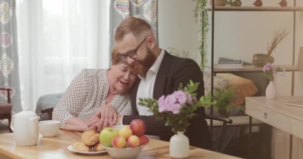 Stilig man i kostym och glasögon spenderar tid med sin mamma hemma. Son och hans mor sitter tillsammans vid bordet, leende och omfamnad.Begreppet omsorg och kärlek. — Stockvideo