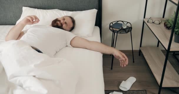 Bonito homem de 30 anos acordando de manhã e parecendo cansado enquanto lia tarde. Um adulto barbudo a dormir na cama e a levantar-se com dores de cabeça, a tocar na cabeça. Conceito de estilo de vida . — Vídeo de Stock