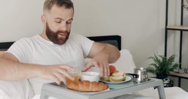 ハンサムな髭を生やした男は、足に食べ物がいっぱいのトレイに座って朝を楽しんでいました。ベッドで朝食をとりながら30代の幸せな男が手をこすりました。リラックスの概念。ホーム背景. — ストック動画