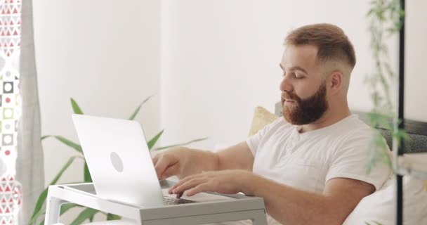 Homem adulto barbudo usando seu computador portátil enquanto fala e digita. Bonito tipo de 30 anos a trabalhar e a ter videoconferência enquanto estava sentado na cama. Conceito de trabalho em casa . — Vídeo de Stock