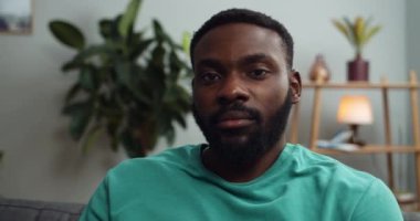 Kanepede oturan ve kameraya poz veren ciddi bir adamın yakın görüntüsü. Kameraya bakan yakışıklı Afro-Amerikalı adamın portresi. İnsanlar ve duygular kavramı. Ev arkaplanı.