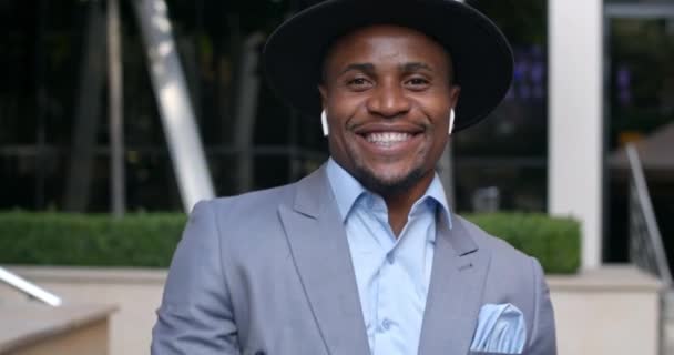 Close-up uitzicht op stijlvolle Afro-Amerikaanse man in hoed en klassiek pak met een goede stemming, bewegend en glimlachend. Knappe man in draadloze hoofdtelefoon dansend en luisterend naar muziek op straat. — Stockvideo