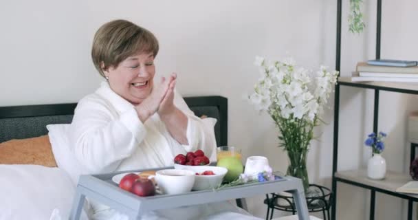 Oude mooie dame zittend met trey op haar benen en genietend van de ochtend. Gelukkig volwassen vrouw wrijven haar handen en op zoek naar camera tijdens het ontbijt op bed. Concept van ontspanning. — Stockvideo