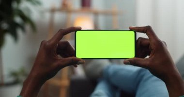 Genç adam kanepede uzanıyor ve yatay manzara modunda yeşil ekran akıllı telefon tutuyor. Adam ekrana bakarken video izliyor. Krom anahtar kavramı. Rahat ev arkaplanı.