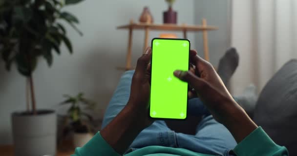 Afro american man scrolling news feed and looking at mock up screen with trecking markers. Guy using mobile phone and browsing internet while lying on sofa. Concept of chroma key. — Stock Video