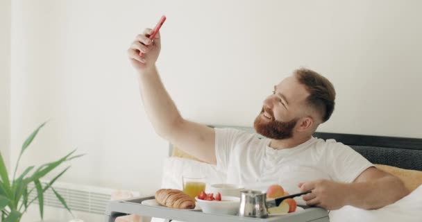 Yakışıklı sakallı adam sabah kahvaltı ederken selfie çekiyor. 30 'lu yaşlarda bir adam akıllı telefonunun ön kamerasına poz veriyor ve bir tepsi dolusu yiyecekle yatakta oturuyor.. — Stok video