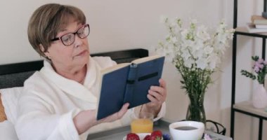 Ayakları yemek dolu bir şekilde otururken emekli bir kadının masal okumasını yakından izleyin. Gözlüklü yaşlı kadın yatakta kahvaltı ederken kitap okuyor. Boş zaman kavramı..