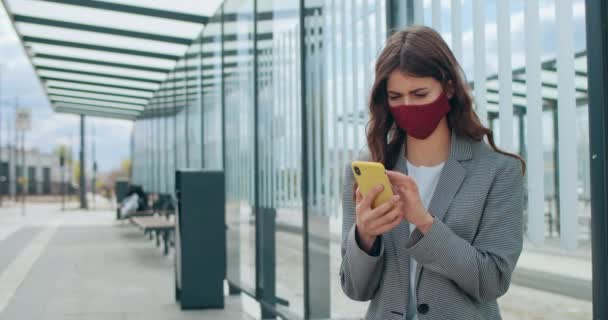 Jonge vrouw in herbruikbaar gezichtsmasker surfen op internet of sociale netwerken. Brunette mooi meisje met behulp van moderne smartphone en scrollen scherm tijdens het zitten bij de bushalte. Concept van COVID. — Stockvideo