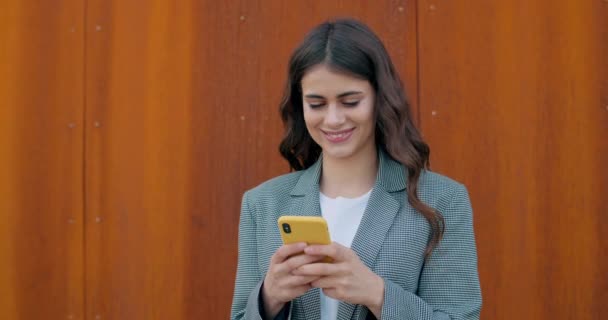 Joven chica morena atractiva sonriendo mientras se utiliza el teléfono inteligente y escribiendo mensaje.Alegre mujer milenaria navegando por Internet o charlando en las redes sociales. Al aire libre . — Vídeos de Stock