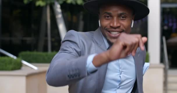 Hombre afro americano alegre en auriculares inalámbricos sonriendo y bailando en la calle. Chico guapo con traje y sombrero escuchando música y moviéndose mientras tiene buen humor . — Vídeo de stock