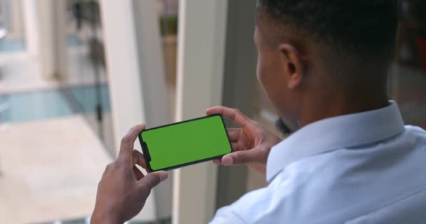 Overschouderbeeld van een Afro-Amerikaanse man met smartphone in horizontale landschapsmodus. Een man die naar een telefoonscherm kijkt. Concept van chroma sleutel. Moderne kantoorgebouw achtergrond. — Stockvideo