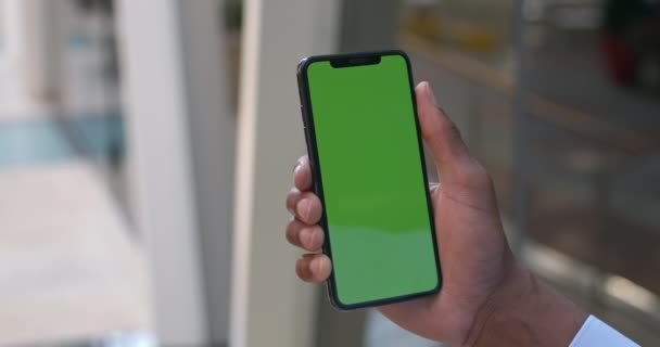 モックアップ画面とスマートフォンを使用して男の手。携帯電話の緑の画面に触れるとズームアフリカ系アメリカ人の男のビューを閉じます。クロマキーの概念。近代的なオフィスビルの背景. — ストック動画