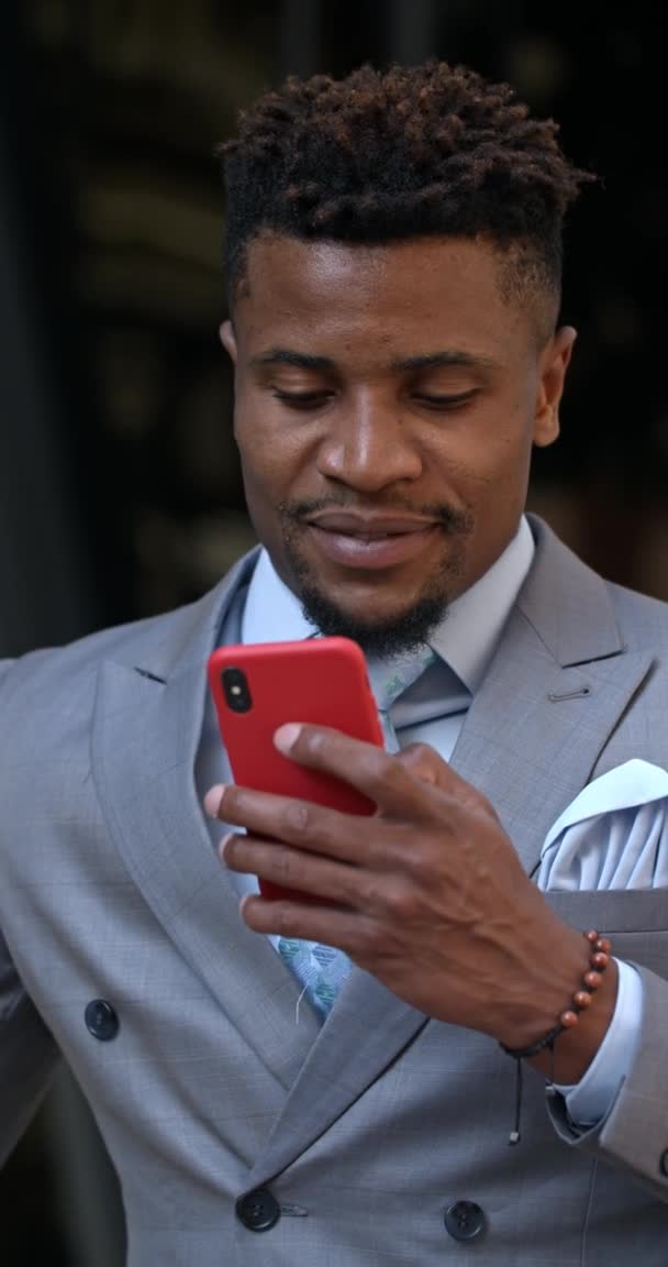 Snygg vuxen man i 30-årsåldern med sin moderna smarttelefon och leende. Framgångsrik kille i kostym och slips meddelanden eller surfa internet medan du tittar på telefonskärmen. Vertikal video. — Stockvideo