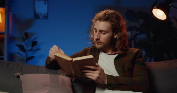 スマートな男がソファに座って、自宅でランプライトで文学を読んでいます。夜の無料時間を楽しみながら本のページをめくる千年男.余暇の概念. — ストック動画