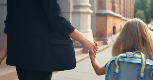 Крупним планом вид маленької блондинки в уніформі з сумкою, що йде з її матір'ю в руці. Вид ззаду на дитину, що тримає маму і вступає в школу рано вранці. Концепція відкритих дверей . — стокове відео