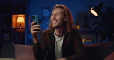 Neşeli milenyum adamı arkadaşlarıyla video sohbeti yaparken el sallıyor ve konuşuyor. Genç adam kanepede oturup gülümserken akıllı telefonu evde iletişim kurmak için kullanıyor..