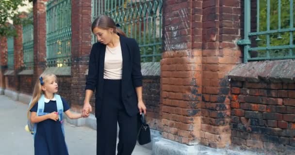 Mãe atraente em terno clássico segurando a mão de sua filha e andando. Jovem mulher conversando com seu filhinho enquanto vai a caminho da escola. Conceito de comícios familiares . — Vídeo de Stock