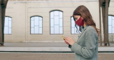 Akıllı telefon kullanan ve yürüyen genç bir kadının yan görüntüsü. Boş tren istasyonunda el yapımı maske takan ve ekrana dokunan bin yıllık esmer kız görüntüsü.